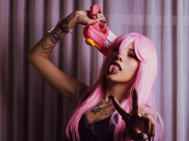 SharlotteX with pink hair, tongue out, holding pink toy, tattooed arm, wearing black top, making "sh" gesture with fingers, looking playful. Her tits are partially visible.