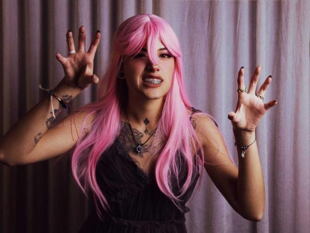SharlotteX with pink hair, black dress, tattoos, and black nail polish, hands up, tongue out, showing metal teeth, wearing rings and bracelets, looking playful and mischievous.