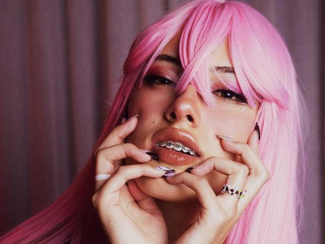 SharlotteX com cabelo rosa, mãos no rosto, mostrando os dentes com aparelho metálico, usando vários anéis, sombra rosa e roxa, unhas longas, olhando sedutivamente para a câmera..