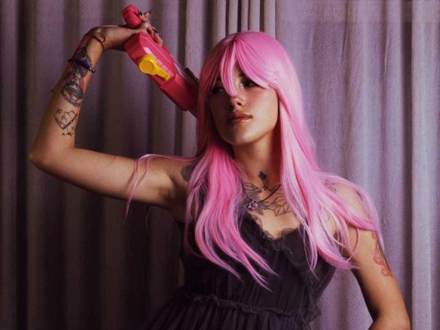 SharlotteX with pink hair, black dress, holding red toy gun, tattoos on arms, raising arm, looking at camera, purple curtain background, slightly parted lips.