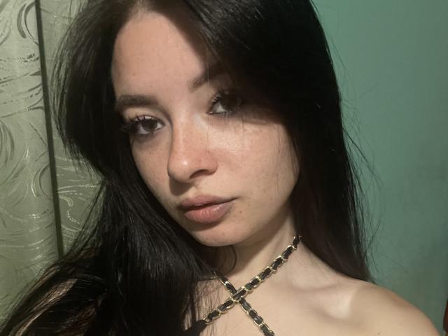 Linok with long black hair, pale skin, and small tits, wearing a gold chain necklace, looking at the camera with a slight pout, green background.