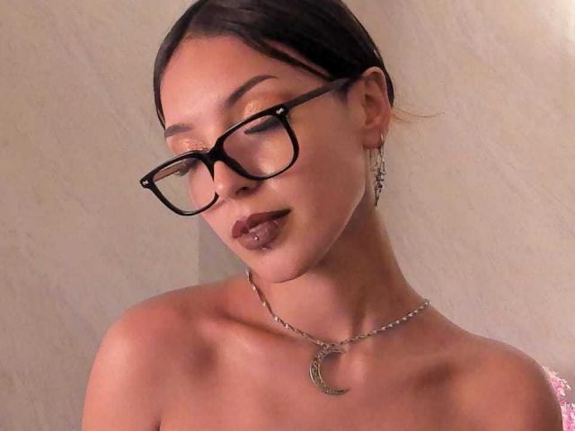 ViolettSoul, con cabello castaño oscuro, gafas y un collar con forma de luna creciente, sin ropa, mostrando sus hombros desnudos y sus labios castaños oscuros con perforaciones, y con aretes colgantes..