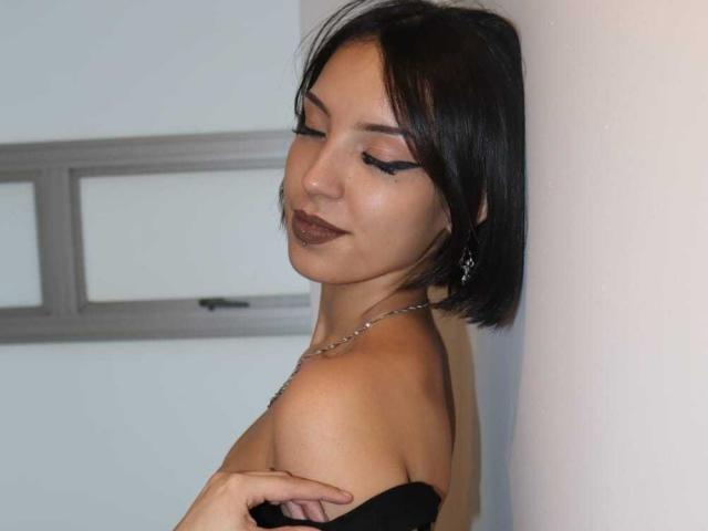 ViolettSoul tiene el cabello corto y negro, lápiz labial oscuro y lleva delineador de ojos negro. Está apoyada contra una pared blanca, tirando de una correa negra, mostrando su hombro desnudo..