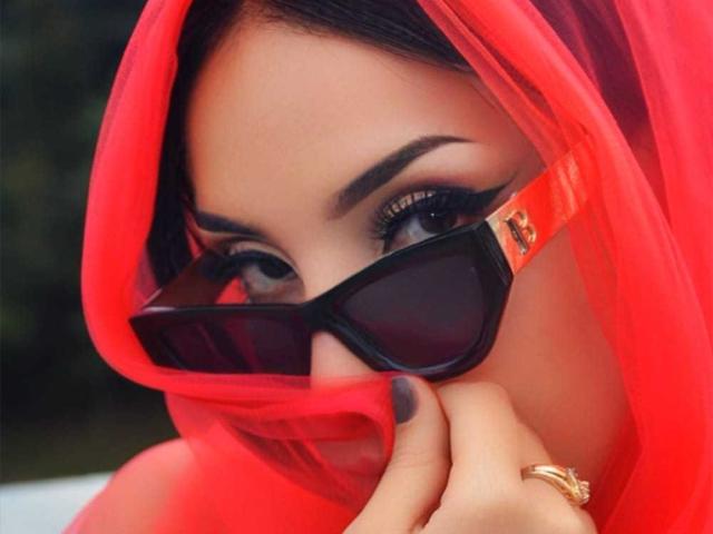ViolettSoul con eyeliner scuro, indossa occhiali da sole neri, un hijab rosso trasparente e un anello dorato, con un dito che tocca le labbra. Le sue unghie sono dipinte di grigio scuro..
