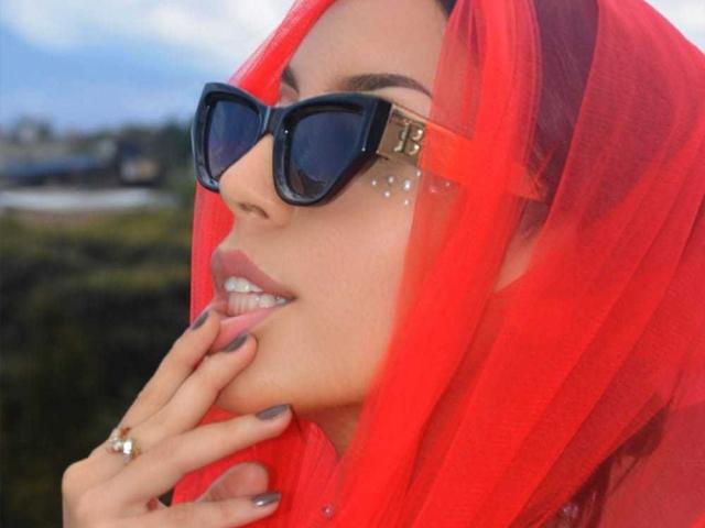 ViolettSoul com um véu vermelho transparente, óculos de sol pretos grandes, piercings de prata no nariz, unhas cinzas e um anel de ouro, mordendo o dedo, sorrindo, mostrando seus dentes brancos..