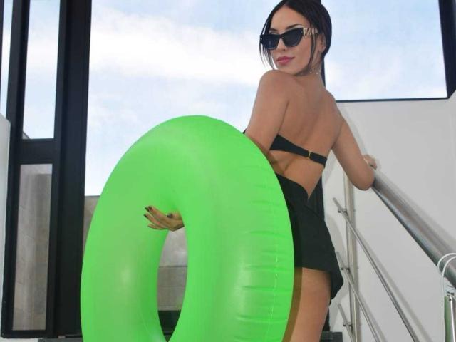 ViolettSoul con cabello oscuro recogido en un moño, vistiendo un bikini negro, sosteniendo un aro inflable verde, de espaldas a la cámara, mirando por encima del hombro, usando gafas de sol, de pie en las escaleras..