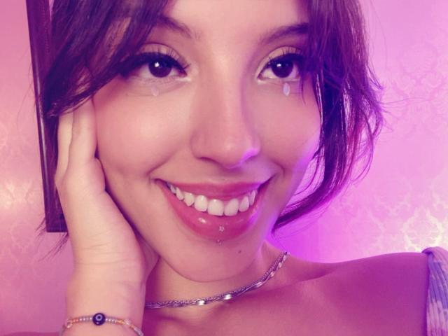ViolettSoul, com um sorriso, olhos castanhos e um piercing no nariz, tocando sua bochecha com a mão, usando um colar e pulseira, rosto iluminado em tons de rosa, mostrando seu lábio inferior com piercing..