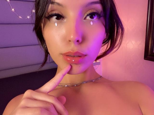ViolettSoul tiene ojos brillantes, labios rosados y llenos con un brillo lustroso, y un dedo tocando su barbilla. Lleva un collar de plata y una iluminación morada-azul..