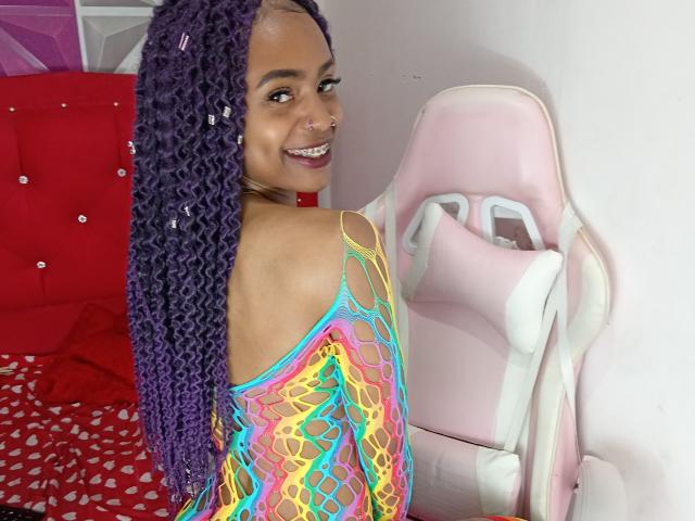 KatalinaCandy with purple braids, smiling, wearing a neon rainbow mesh top, showing her back, nose ring, and looking over her shoulder, sitting on a pink gaming chair.