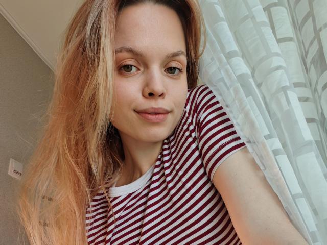Sinteas, avec ses longs cheveux blonds, ses yeux verts et un léger sourire, porte une chemise rayée rouge et blanche et se tient près d'un rideau blanc. Ses seins sont petits et elle est mignonne..