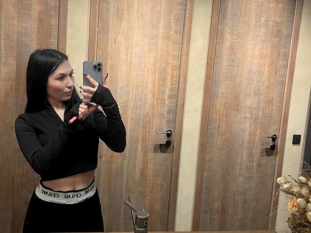 MarianneChristiana with long black hair, black crop top, black pants with "PUMA" waistband, taking a mirror selfie in a wooden room, holding a silver iPhone.