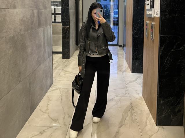 MarianneChristiana with black leather jacket, black pants, white heels, holding phone to face, black bag in hand, standing in marble hallway, taking selfie.
