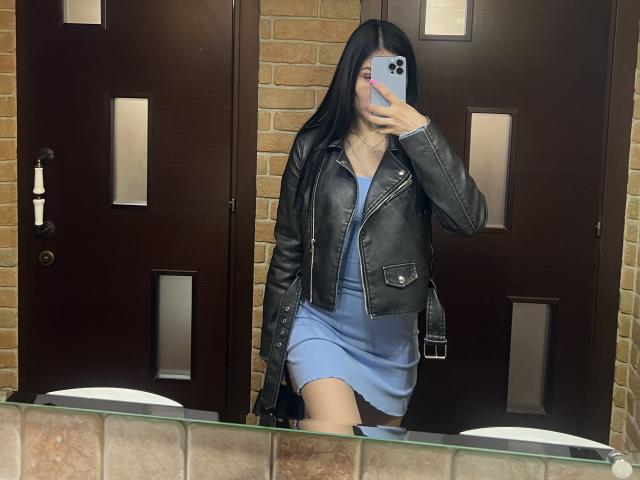 MarianneChristiana with long black hair, wearing a blue dress and black leather jacket, taking a mirror selfie, showing her ass, holding a phone, in a bathroom.