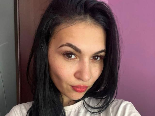 MarianneChristiana has black hair, brown eyes, and red lips with a nose ring. She's wearing a white shirt, looking slightly down with a subtle smile.