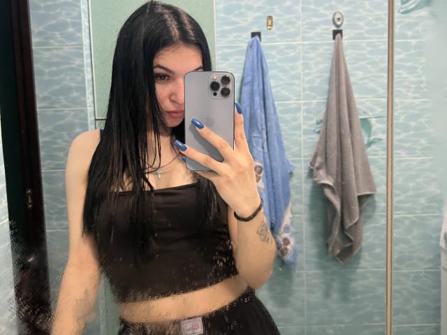 MarianneChristiana with long black hair, blue nails, black crop top, and black skirt, taking a mirror selfie in a teal-tiled bathroom, showing her small tits and tattoo on her arm.