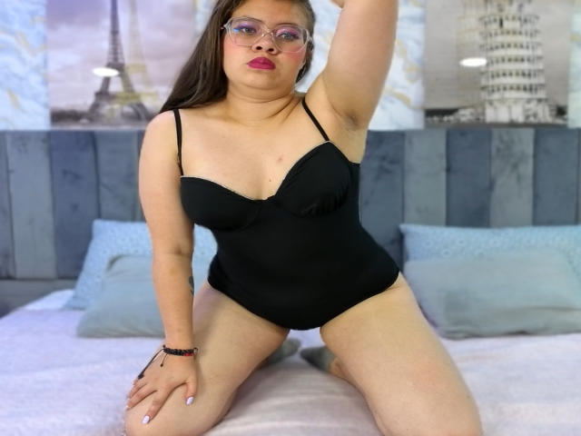 Isabella Reid con gli occhiali, che indossa un costume intero nero, inginocchiata su un letto, con un braccio alzato, che mostra i suoi seni, la vulva appena visibile, rossetto rosso, braccialetto al polso sinistro..