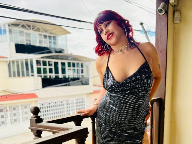 AnabellaThompson with red hair, wearing a sparkly black dress, showing her tits, standing by a balcony, smiling, red lipstick, choker, earrings, hand on railing, daytime.