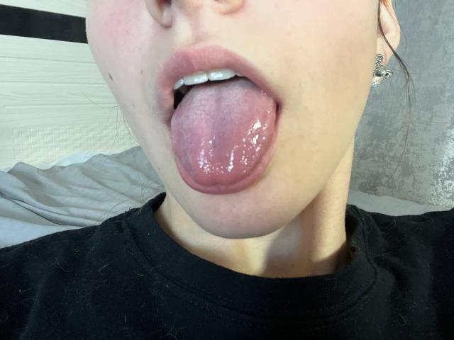 Das Gesicht von VinnaSkyHott, Zunge heraus, glänzend rosa mit Speichel, Lippen geöffnet, weiße Zähne sichtbar, trägt ein schwarzes Hemd, kleine Nase, helle Haut, Ohr mit silbernem Ring, Bett im Hintergrund..