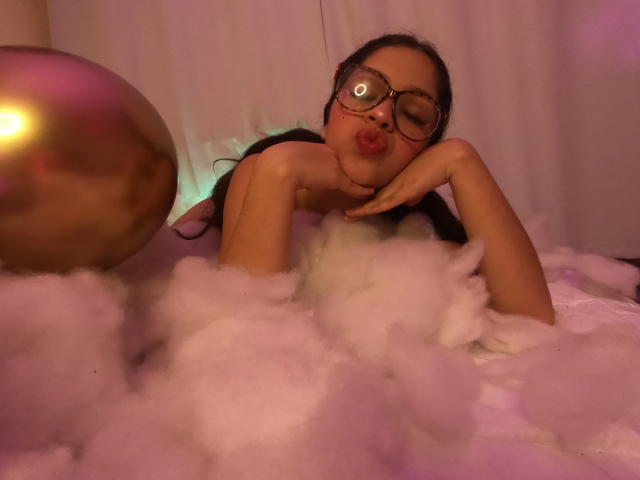 NatashaSteven with pouty lips, wearing glasses, and lying in fluffy pink foam, her tits and ass partially covered, with a shiny gold ball to the left.