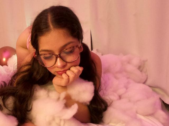 NatashaSteven with glasses, long brown hair, pink bra, and puffy white foam, on knees, head resting on hand, looking down, pink and white lighting, soft focus.