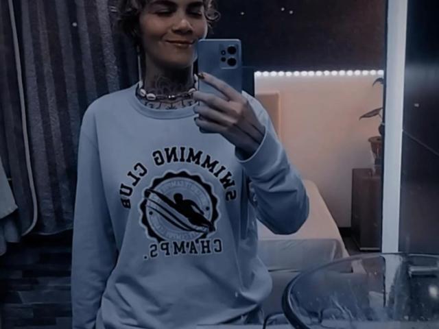 AnaRoa, con i capelli castano ricci e corti, sorride, indossa una felpa azzurra "Soul Swimwear", tiene in mano un telefono blu, ha un tatuaggio visibile sul collo, ed è in piedi in una stanza poco illuminata..