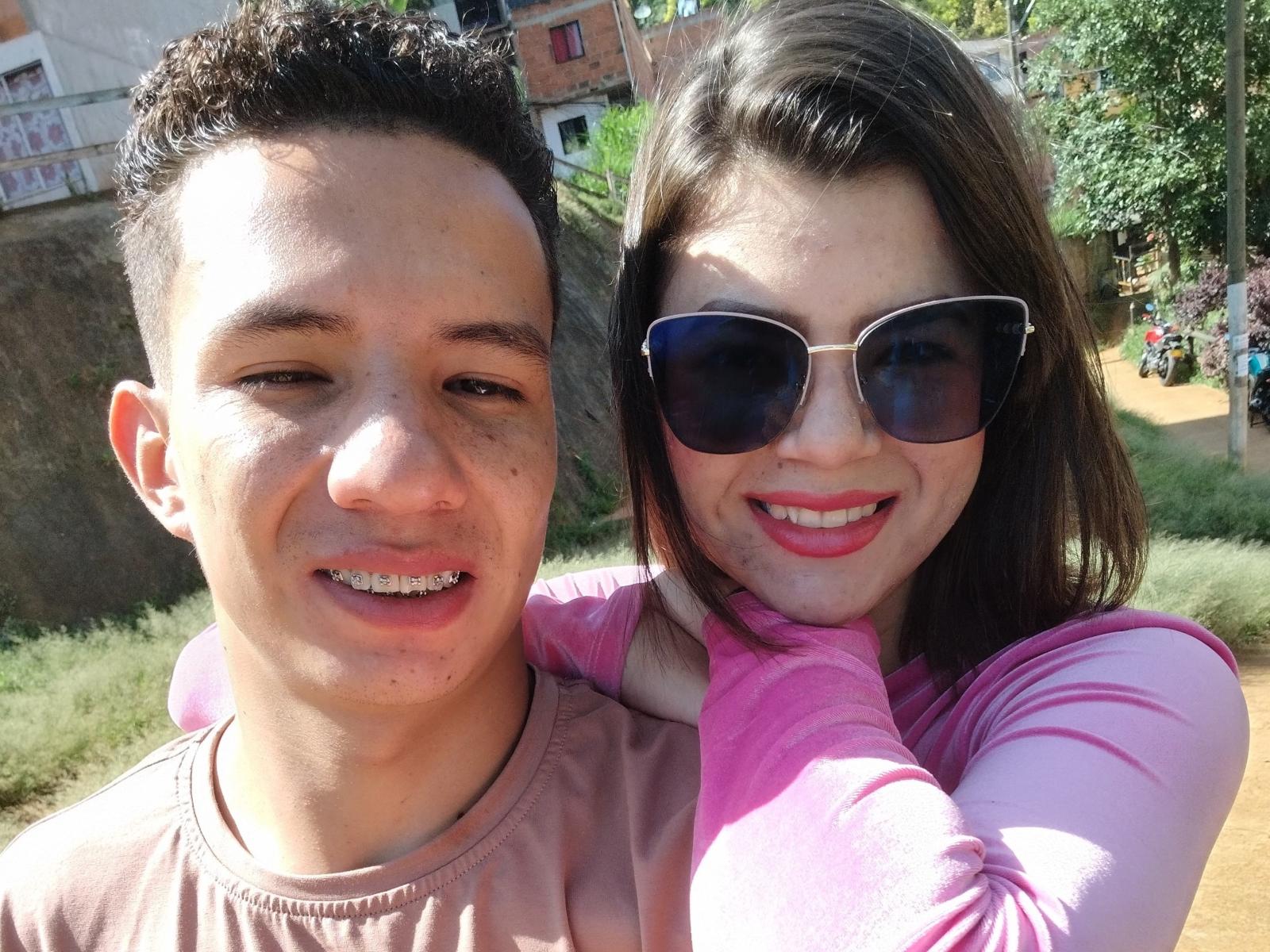 SabrinaJeremy has a cute face with pink lips and brown hair, wearing sunglasses and a pink shirt. She's hugging a guy with short curly hair and a pink shirt.