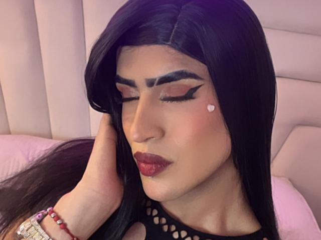 ScarlettTs with dark, straight hair, closed eyes, bold winged eyeliner, pink heart on cheek, red lipstick, black fishnet top, hand touching head, pink bed in background.