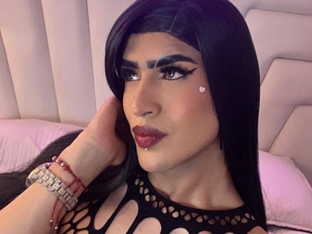ScarlettTs with long black hair, bold eyebrows, and dark lipstick, wearing a black fishnet top, a heart tattoo on the cheek, and a bracelet with red beads.