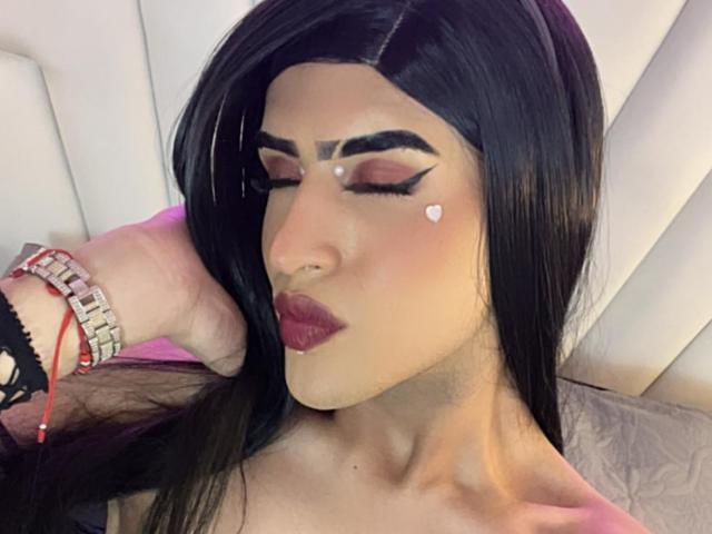 ScarlettTs with long black hair, bold eyeliner, heart-shaped cutout on cheek, pink lipstick, hand on head, wrist bracelet, and nose piercing, looking down.