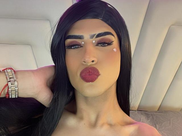 ScarlettTs with long black hair, dark eyebrows, red lipstick, and small white dots on her face, pouting with a hand on her head, wearing a silver bracelet.