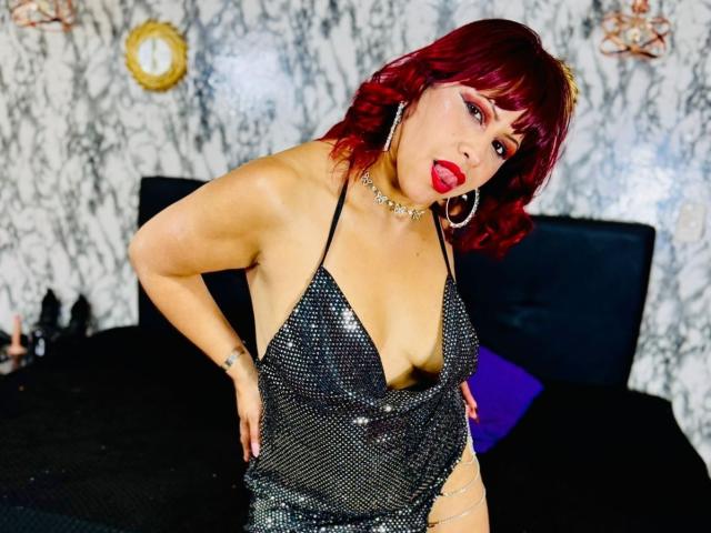 AnabellaThompson with red hair, red lipstick, and large hoop earrings, wearing a sparkly black halter dress, exposing her boobs, and a chain on her hip, posing seductively.