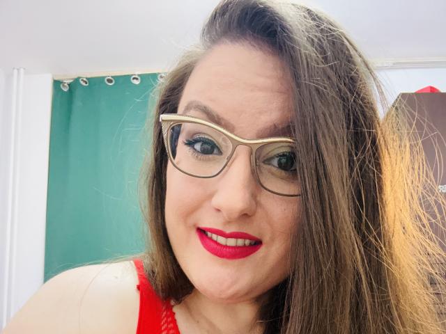 BadAngel with long brown hair, glasses, and red lipstick, smiling at the camera, wearing a red lace top, showing off her tits. Her pussy isn't visible.