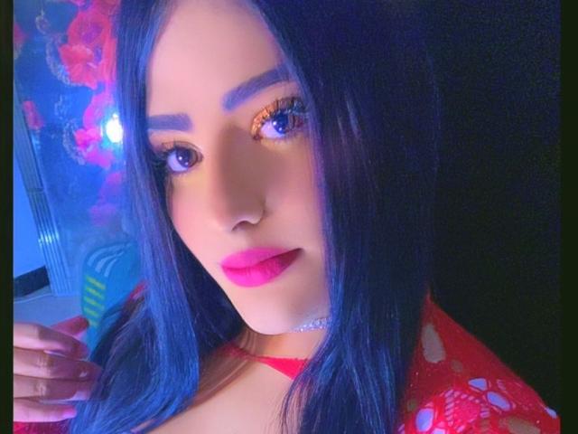 SexyHottLatinaX with blue hair, purple eyes, pink lips, red top, and a nose ring, looking at the camera, holding her hair with one hand.