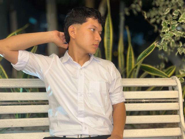 LukaMiranda, with short black hair, wearing a white button-up shirt, sits on a white bench, right arm raised, looking to the side with a neutral expression.