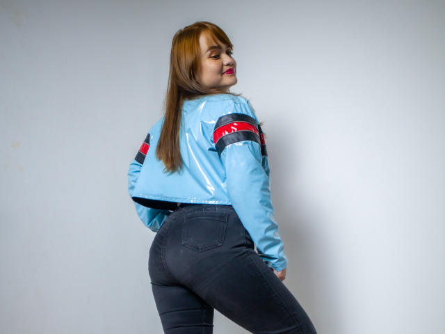 MoonRay69 tiene una chaqueta azul claro brillante, brazaletes rojos y negros, jeans negros, y está de espaldas, mostrando su trasero. Tiene el pelo largo y rojo y una sonrisa linda..