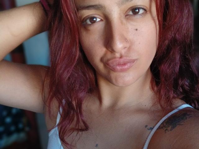 LyllaStar has red, wavy hair, a nose piercing, and a tattoo on her right shoulder. She's wearing a white tank top, showing small tits. She's looking at the camera.