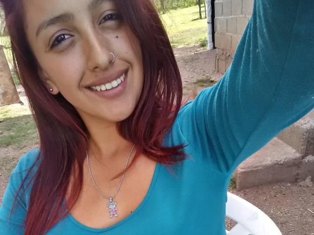 LyllaStar with red hair, smiling, nose ring, and blue top, showing off her tits with a pink pendant necklace in a selfie.