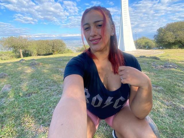 LyllaStar with red hair, medium tits in a black tee, pink shorts, crouching, smiling, holding tee, outdoor, grassy, tall white monument in background, blue sky, fluffy clouds.