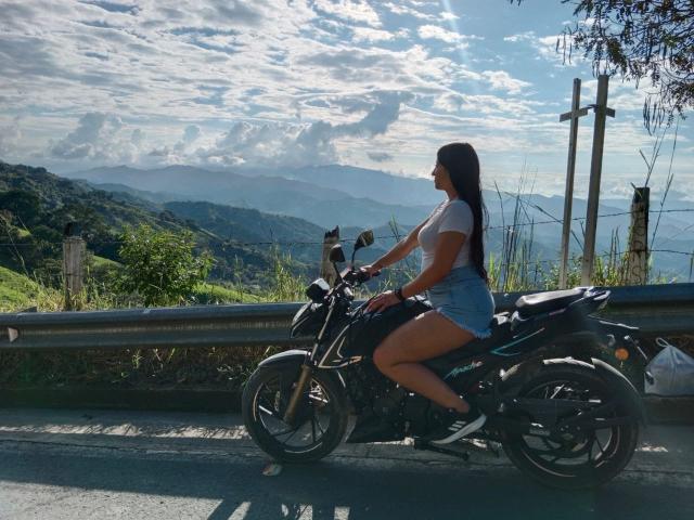 ZaraBeicker with long black hair, white t-shirt, and denim shorts, riding a black motorcycle on a sunny road, mountains and cloudy sky in the background.