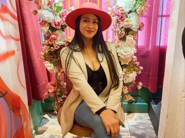 ZaraBeicker, with long black hair, wearing a pink hat, beige coat, black lace top, and blue jeans, sitting on a wooden stool, smiling, surrounded by pink and white flowers.