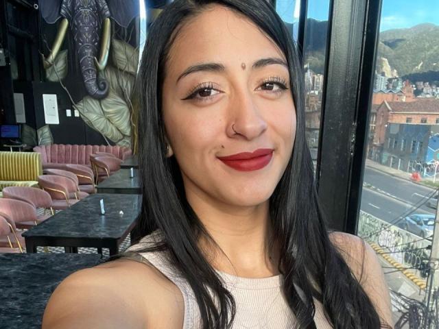 ZaraBeicker, with dark hair, red lipstick, and a nose ring, smiling at the camera, showing her tits in a sleeveless top, in a cafe with a window view.