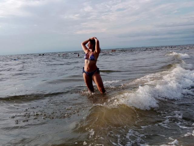 Zara Beicker, vestindo um biquíni azul, está com os joelhos na água do oceano, as mãos na cabeça, com ondas ao redor, e um céu nublado acima, com um horizonte distante ao fundo..