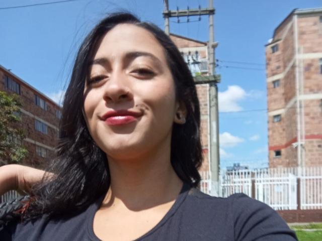 ZaraBeicker tiene el cabello oscuro, piel de color marrón medio y está sonriendo con labios rojos, vistiendo una blusa negra, en un entorno exterior soleado con un cielo azul claro..