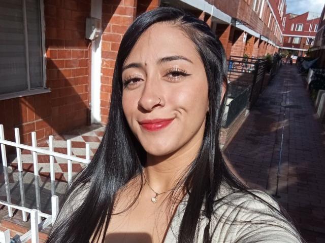 ZaraBeicker has long black hair, red lipstick, and a small nose ring. She's smiling with a cute face and wearing a gray top. Her tits are small, visible.