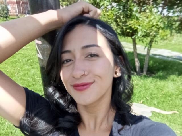 Zara Beicker, con cabello negro ondulado, aro en la nariz, lápiz labial rojo, sonriendo, con la mano en el cabello, vistiendo una camisa negra, hierba verde, un árbol y un poste de madera en el fondo..