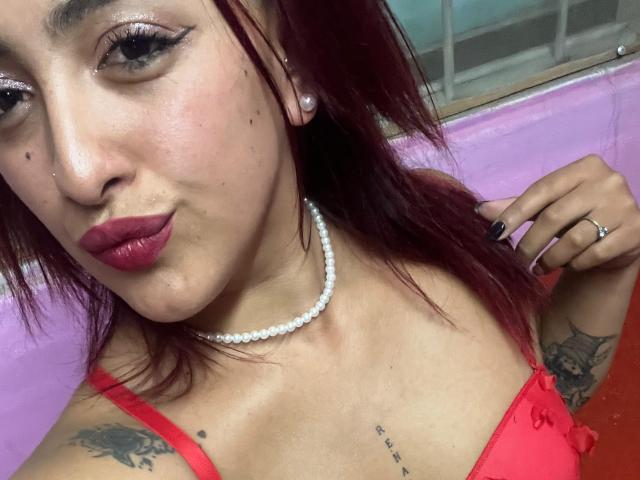 LyllaStar with red lipstick, pearl necklace, red bra, tattoos, and dark nails, pouting, showing her tits, with pink wall and window in the background.