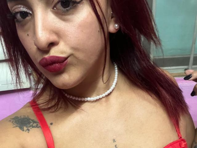 LyllaStar with red lipstick, pearl necklace, and red bra, pouting her plump lips, showing tattoos on her chest and shoulder, with red hair and a nose ring.