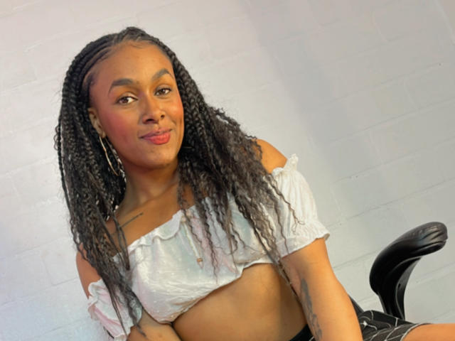 JenaStarr with long braids, wearing a white off-shoulder crop top, showing her tits, and a black chair, smiling with red lipstick, and a tattoo on her left arm.