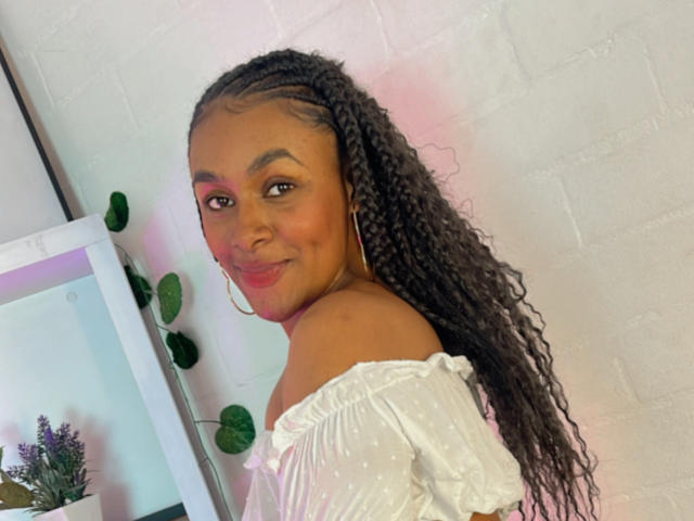 JenaStarr with long, curly black hair, wearing a white off-shoulder top, smiling, pink eye makeup, and large hoop earrings, standing near a plant and mirror.