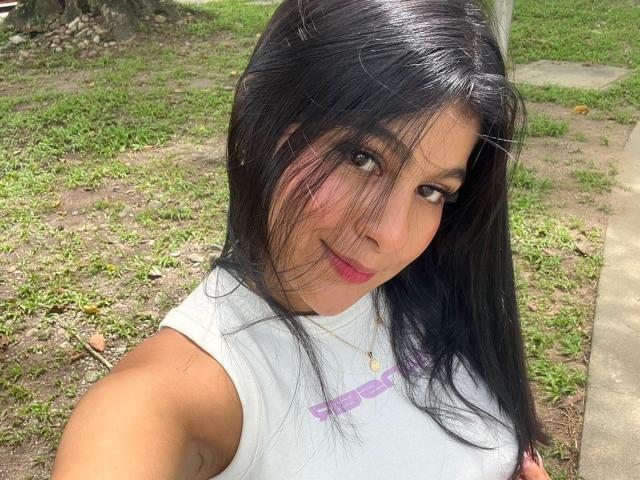 SweetSoffiee has long black hair, brown eyes, pink lipstick, and is wearing a white sleeveless top, showing a slight smile with a gold necklace, outdoors on a sunny day.